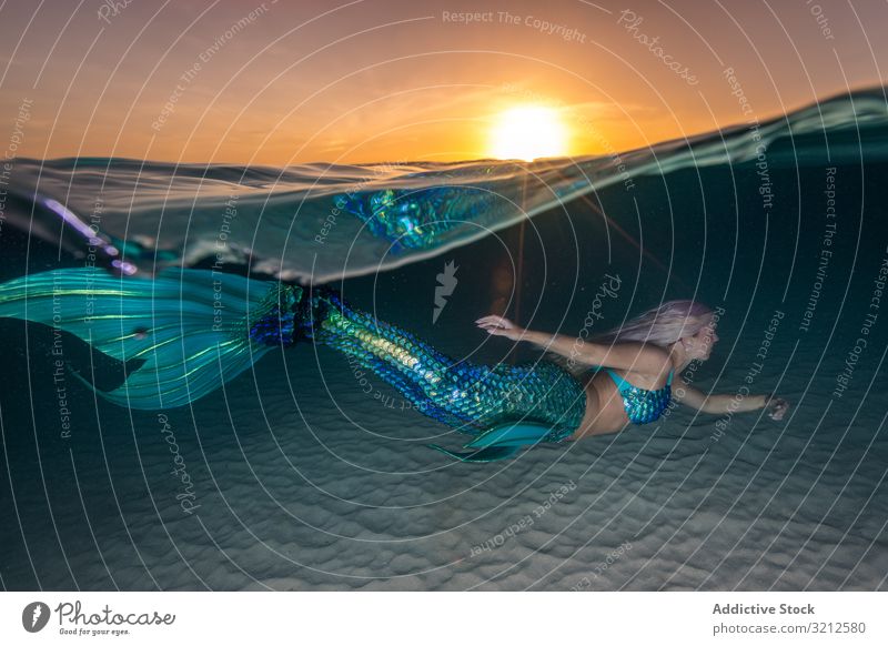 Blond fairytale female mermaid swimming along sea bottom underwater carefree fishtail blue deep blond beautiful daylight woman long hair shiny sunset sky wave