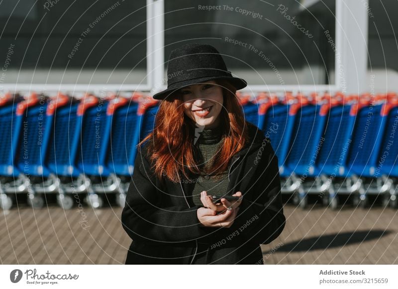 Smiling woman with smartphone in parking lot with shopping carts attractive young beautiful casual cheerful smiling network telephone modern standing joy