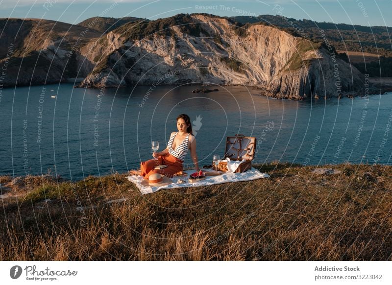 Woman with glass of drink on picnic mat looking at sea and mountains woman seaside beach summer reading leisure sky relax vacation summertime vintage fashion