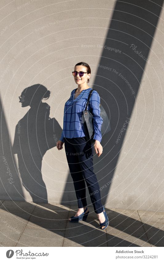 Smiling businesswoman standing near gray wall street smile city building pavement elegant modern urban female manager entrepreneur career bag sunglasses shades