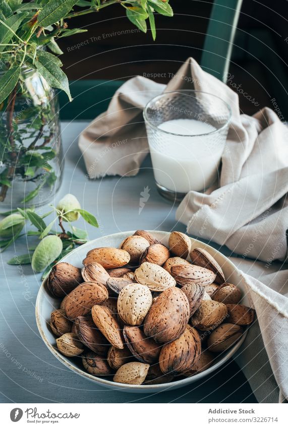 Glass of almond milk next to plate of almonds in shells on kitchen table glass nuts bowl cloth twig green plant rustic snack breakfast ingredient vegetarian