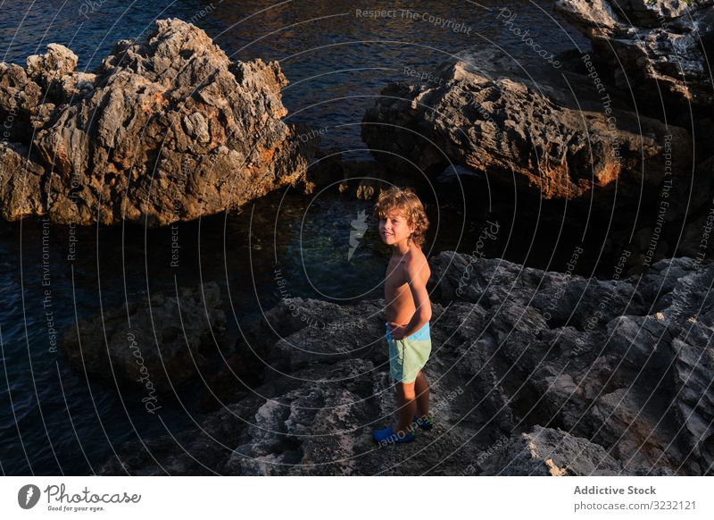 Happy little boy standing on rock at seashore cliff smile wave happy adventure excited seaside carefree coast kid sunset scenic travel delighted beautiful