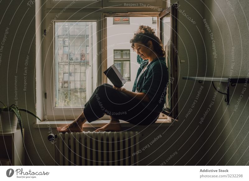 Interested woman reading book at window sill at home interested relaxed literature windowsill barefoot casual sit apartment happy resting young adult peaceful