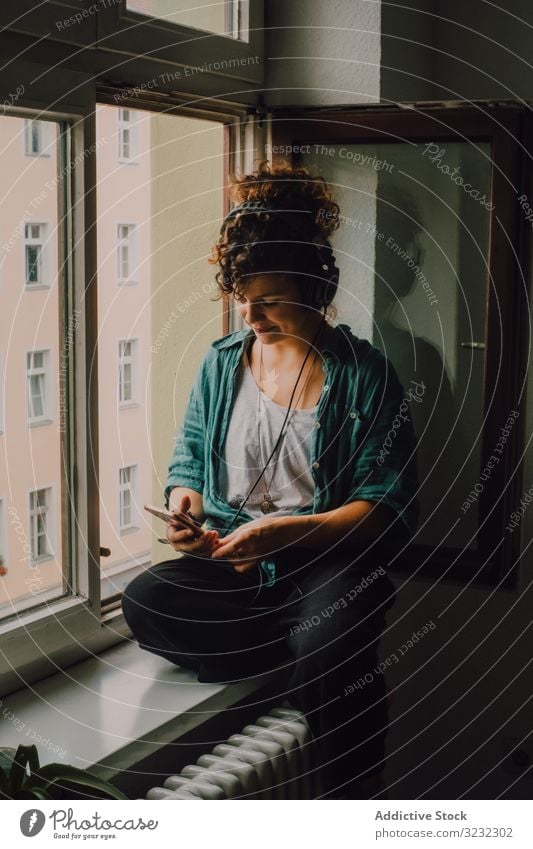 Cheerful woman in headphones using smartphone at home cheerful smile listen happy music browsing window sill curly attractive peaceful relaxed apartment young