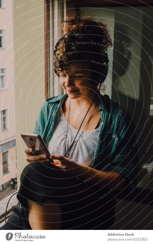 Cheerful woman in headphones using smartphone at home cheerful smile listen happy music browsing window sill curly attractive peaceful relaxed apartment young