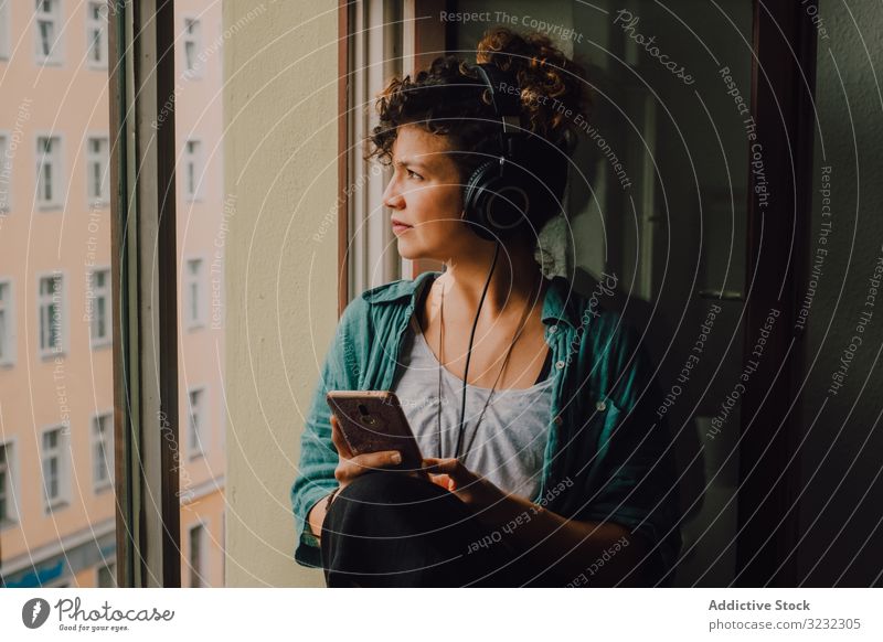 Relaxed woman in headphones using smartphone at home smile pensive listen happy music browsing window sill curly attractive peaceful relaxed apartment young