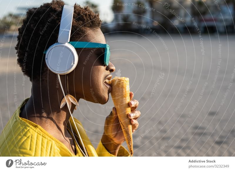 Cool female eating ice cream on beach woman trendy cool desert african american headphones jacket yellow bright sunglasses enjoy sweet food summer fun stick