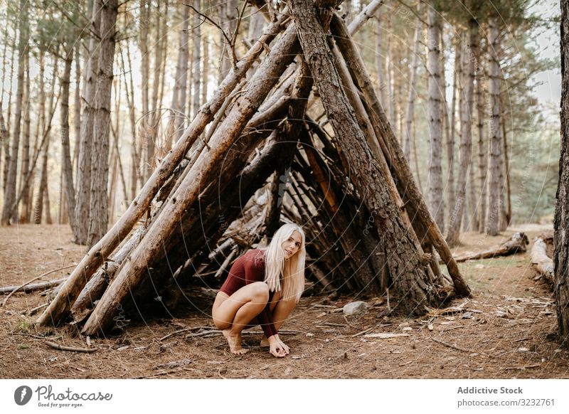 Barefoot woman squatting beside primitive shelter in woods autumn harmony relaxed concept blonde carefree beautiful attractive ecology barefoot trail leotard