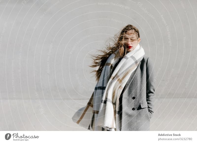 Young woman in coat and scarf on windy day stylish street closed eyes wall city building female urban fashion cool young model outfit warm weather exterior lady