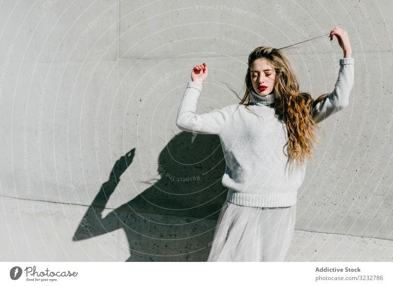 Trendy lady touching hair on street woman stylish sensual closed eyes long hair elegant wall building female model sunny daytime young fashion cool urban outfit