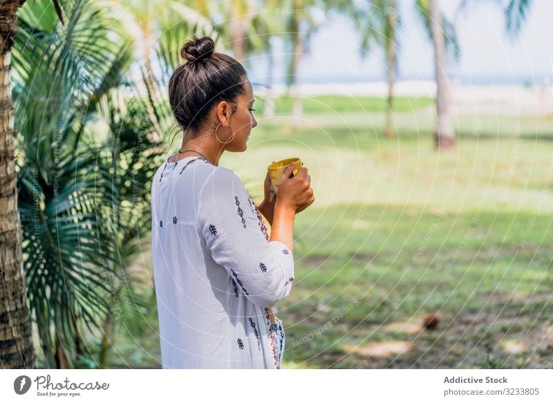 Relaxed woman enjoying hot drink and standing on exotic lawn relaxed seaside palm tree sunny peaceful coffee mug grass tropical rest costa rica young tanned