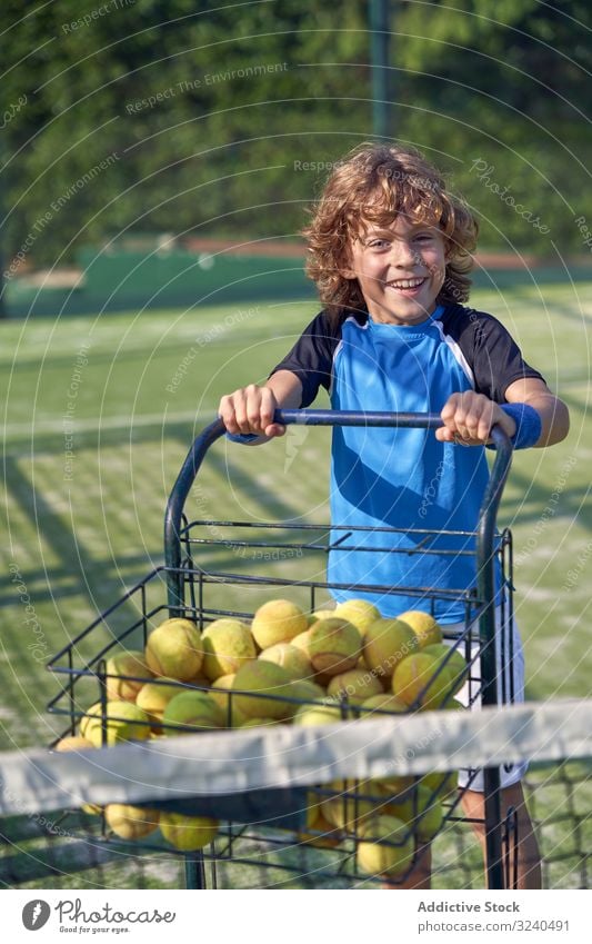 Cheerful boy pushing cart with tennis balls court smile assistant training walk kid child activity game player sport lifestyle practice equipment trolley