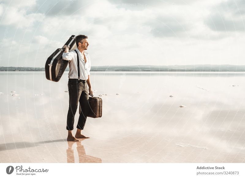 Focused stylish man with suitcase and guitar gig bag on seaside serious barefoot carry water usa shore gazing contemplation modern artist musician young adult