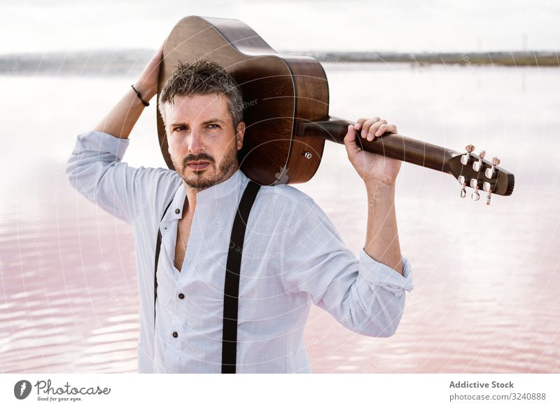 Musician gazing and carrying guitar behind back while standing on beach musician gaze man acoustic modern hold shore young adult white shirt waterfront