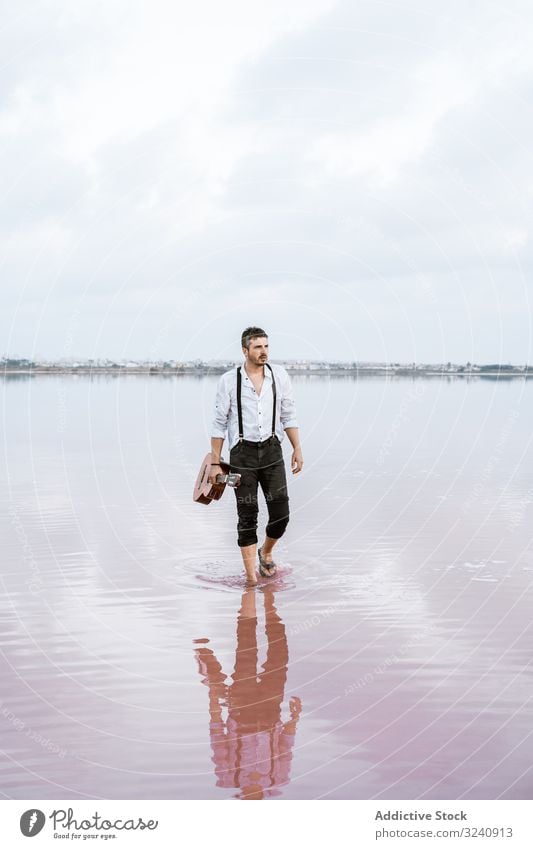 Musician holding guitar at seaside musician inspired barefoot stand play water cloudy man shore white shirt suspenders horizon reflection young guitarist