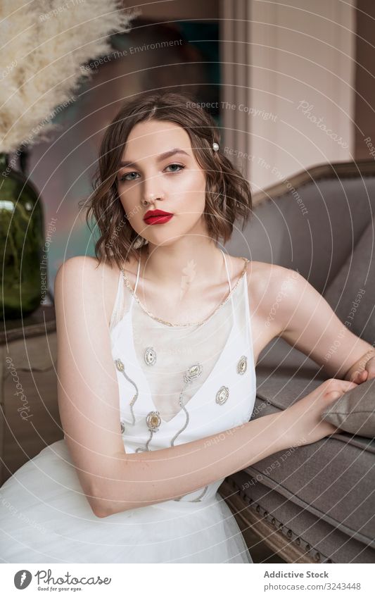 Gorgeous woman with red lips in white dress looking at camera while sitting on floor beside sofa wedding dress vintage chic elegant bride beauty trendy makeup