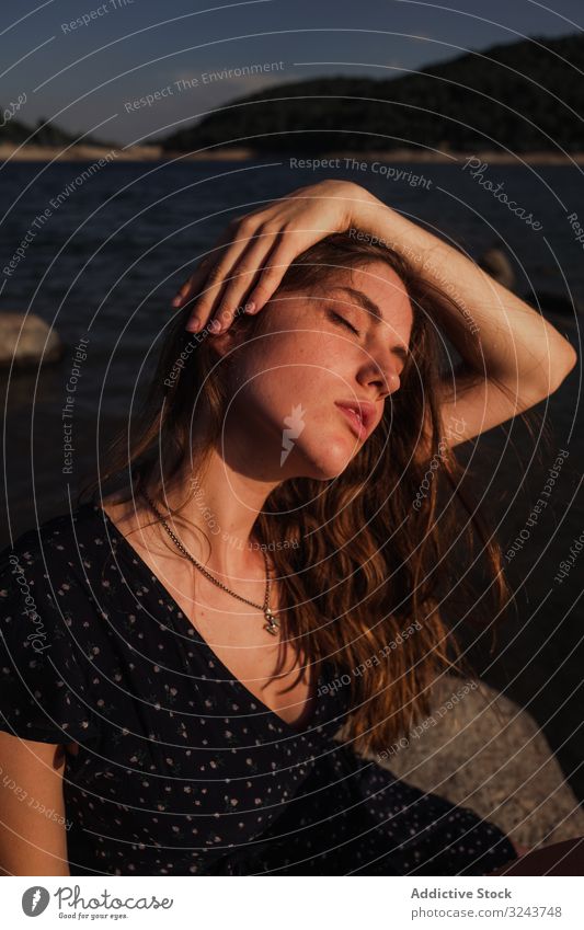 Sensual woman resting on lake shore coast rock touching head sensual summer young dreamy cloudy sky female water vacation weekend lifestyle nature countryside