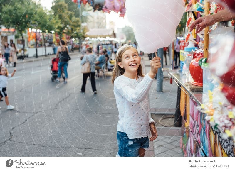 Cheerful girl eating cotton candy on street candyfloss stall smile city fair joy kid happy child urban town sweet fun vendor buy sell sidewalk pavement casual