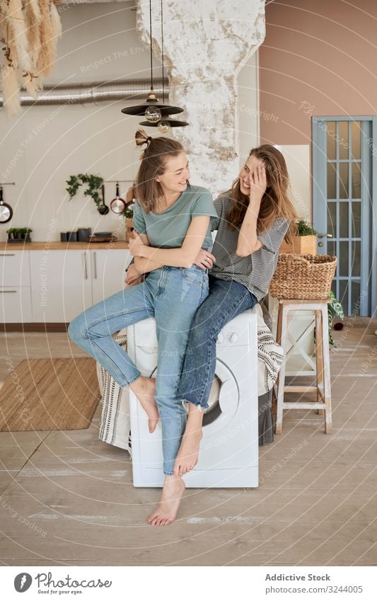 Laughing girlfriends sitting on washing machine in house home fun laugh couple domestic women leisure relocation love lounge barefoot together comfort housework