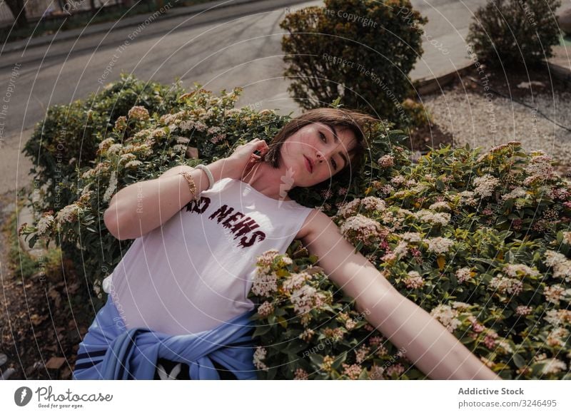 Young lady in trendy casual outfit leaning on shrub with flowers at city street teenager fashionable stylish summer emotionless bloomy hipster youth lifestyle