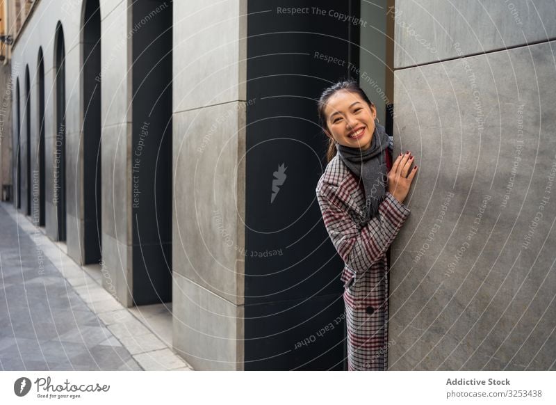 Joyful Asian resting female in casual clothing looking out modern wall of building at city street woman travel town tourist door peer peek playful exploring fun