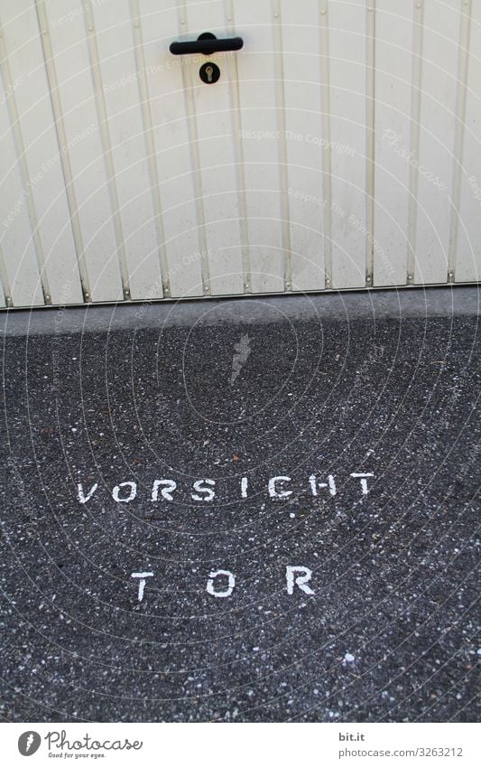 Writing: Caution gate, stands for safety, warning on the floor of the parking lot, in front of a garage with white sheet metal gate with handle. Warning for car, bicycle, pedestrian, against accident, danger, collision with garage door, when opening or closing the door.