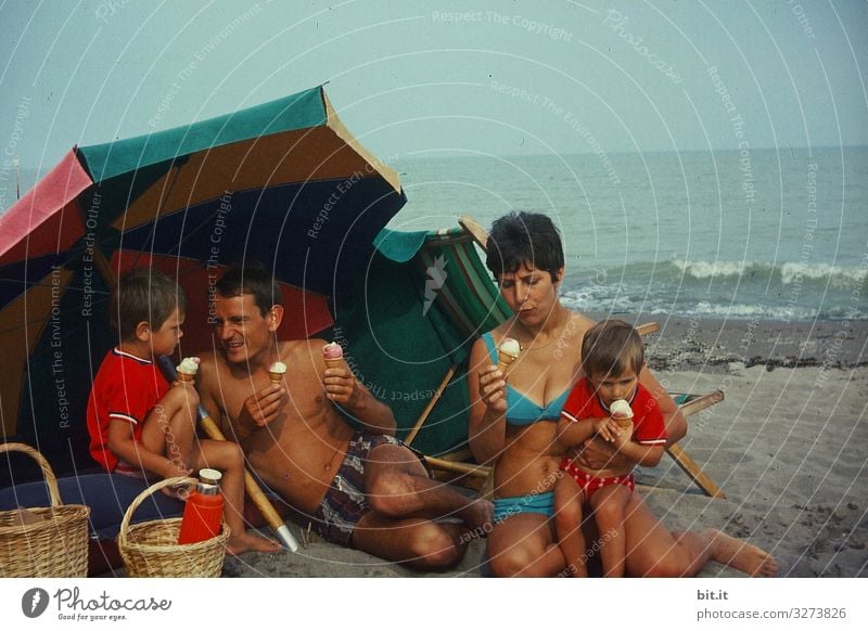 Young, happy family in swimsuits is sitting on the beach, under a parasol, having a picnic with ice cream, in Italy by the sea, back in the 60's. They hold waffle ice cream in their hands and enjoy, eat it in front of the sea.