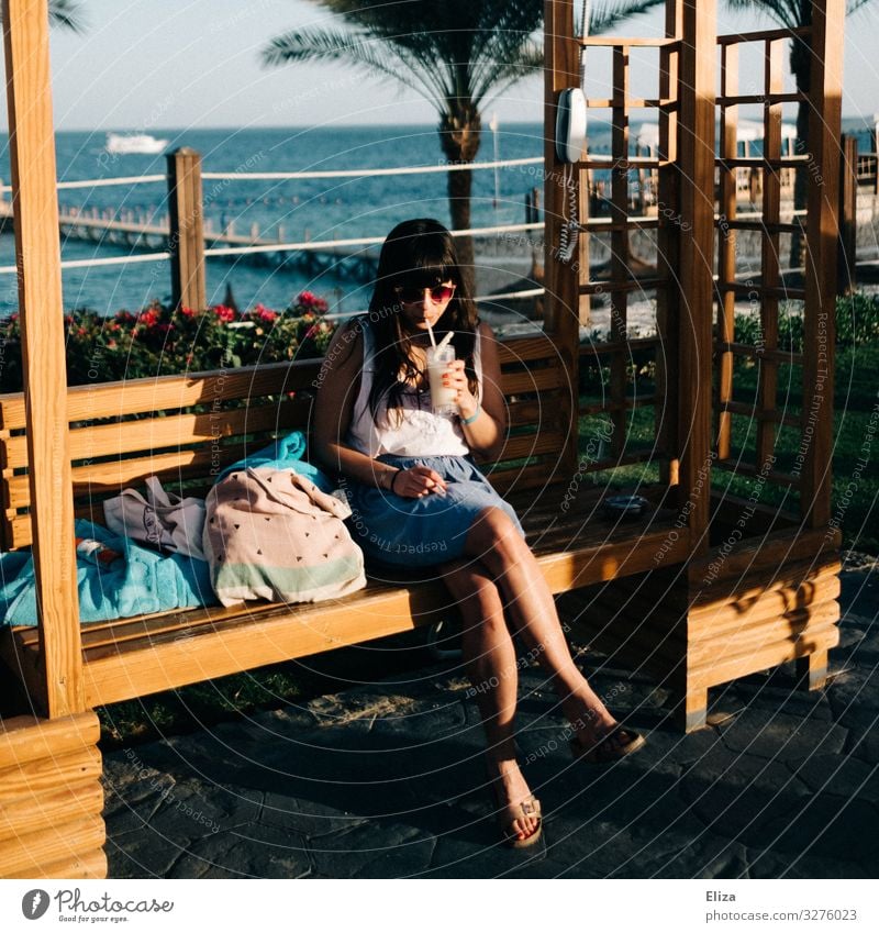 A young woman sitting on a bench in the sun and drinking a drink while on holiday by the sea Feminine Young woman Youth (Young adults) Woman Adults