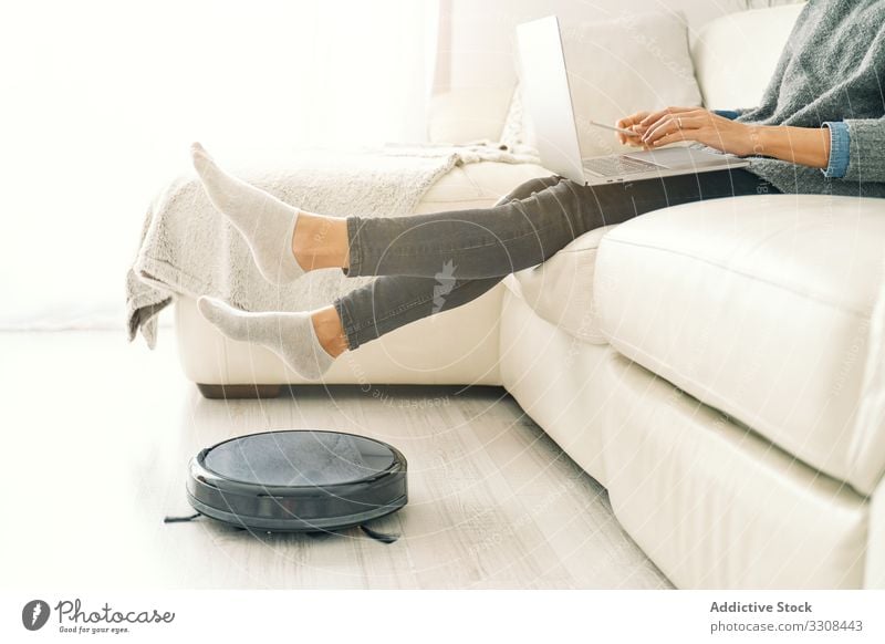 Woman sitting on sofa with feet on robotic vacuum cleaner woman smart home technology floor wireless appliance machine household domestic modern contemporary