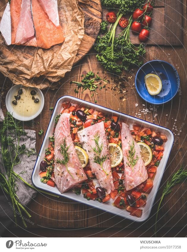 Cooking preparation of Bass fish fillets in Mediterranean sauce with tomatoes, olives and capers in baking pan on rustic wooden background with ingredients. Top view