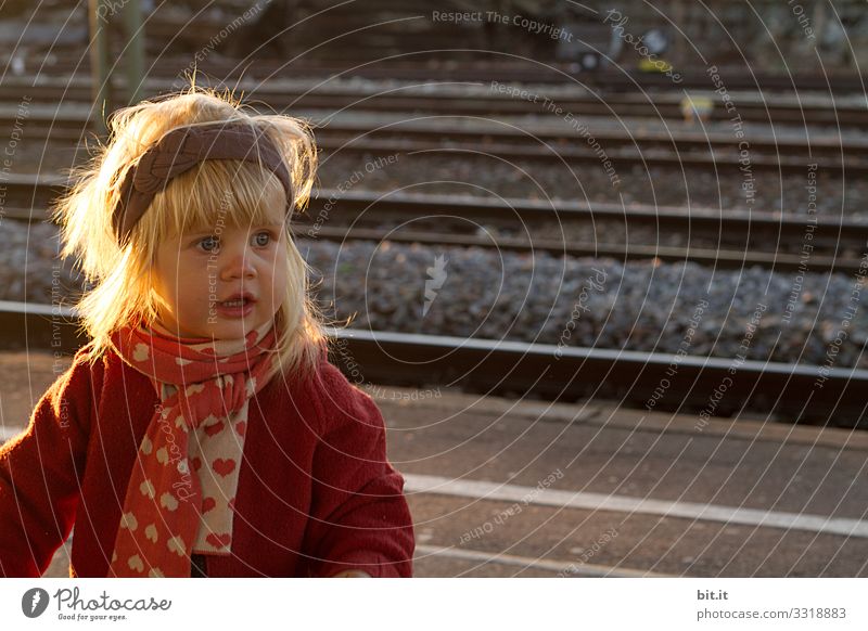 Blonde girl with big eyes, headband & scarf, stands in the autumnal soft evening light at the railway track and waits for visitors. Curious, cheerful child in warm clothes, waiting in the evening sunlight on the platform. Travel, travel by train, train.