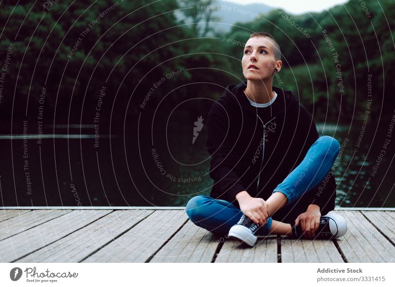 Thoughtful young woman on pier of pond hipster nature short hair piercing casual skinhead female lonely sitting alone green plant tree summer journey wooden