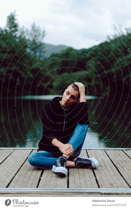 Thoughtful young woman on pier of pond hipster nature short hair piercing casual skinhead female lonely sitting alone green plant tree summer journey wooden