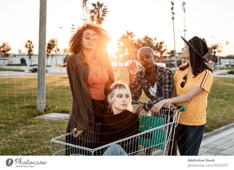 Multiracial group of young women standing around shopping cart on road diverse casual challenge joke empty holding sitting friend team buyer consumerism store