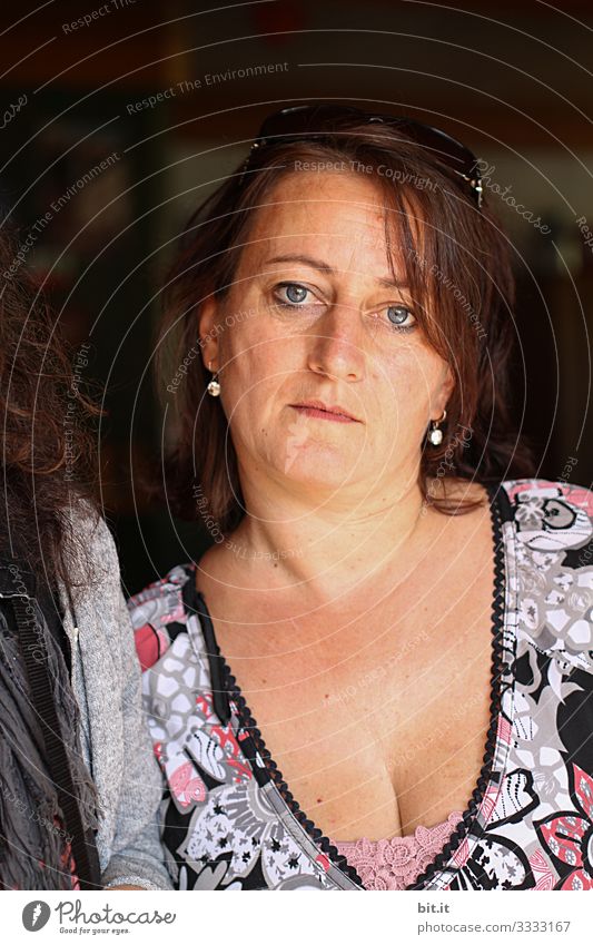 Mature, pretty, modern, beautiful, elegant woman with a low-cut décolleté from a flower dress, is on holiday, in the shade of the house entrance and looks into the camera tired, sad, worried, tired out. The sunglasses, is on her head.