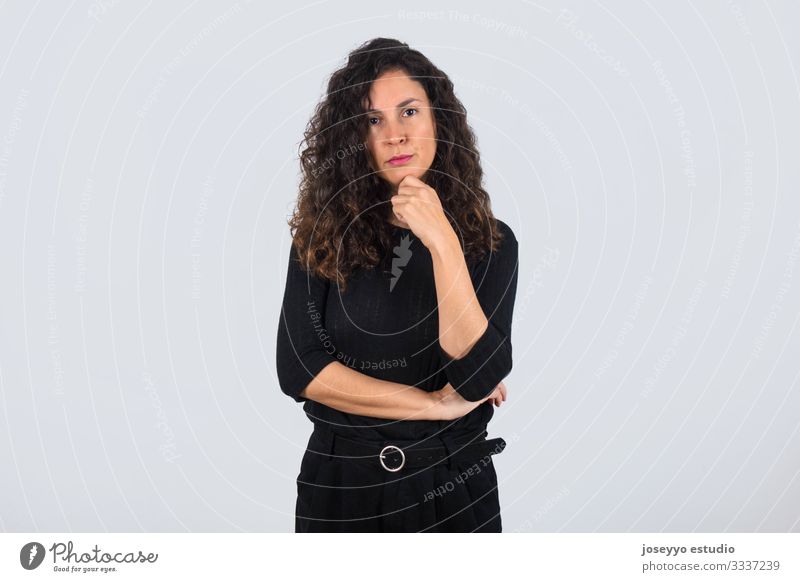 Curly-haired brunette woman dressed in black with her hand on her chin looking at camera with serious expression. 30-40 years advertisement attitude background