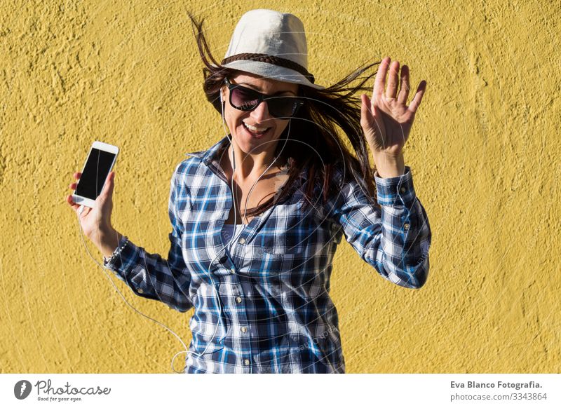 happy beautiful young woman listening to music and having fun over yellow background. She is wearing hat and modern sunglasses. lifestyle Portrait photograph