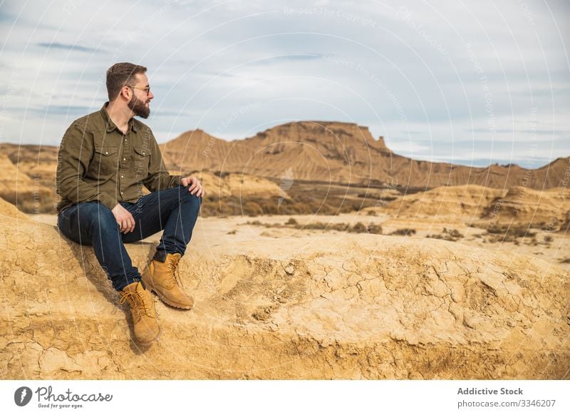 Young man on vacation sitting near cliff at desert tourism mountain rock stone casual sunglasses stylish blue sky smile enjoy travel summer trip nature