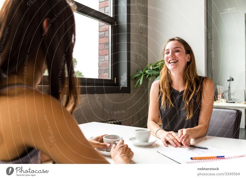 Two young female colleagues discussing work over coffee at the desk. Medium shot. Coffee Happy Desk Table Work and employment Profession Workplace Office