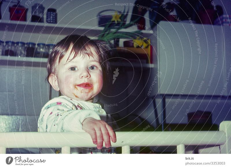 Little girl in her 60s, standing in the playpen in the kitchen, smiling, cheeky, happy, cheerful, mischievously into the camera. Child Toddler Girl Joy Sweet