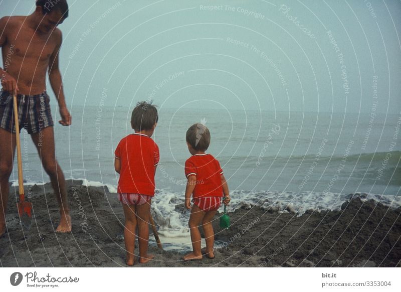 Father with two, small children in swimming trunks and red T-shirts, are standing at the sea and watching a wave of water, which washes into their sand castle on the beach. The family holds shovel and tools in their hands and is curiously amazed by the water play.