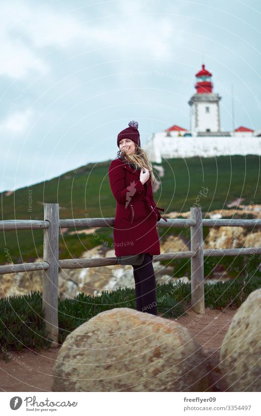 Young coman in a red coat enjoying the wind with a lighthouse on the background woman outdoor winter vertical tourist tourism hiking green grass shoreline