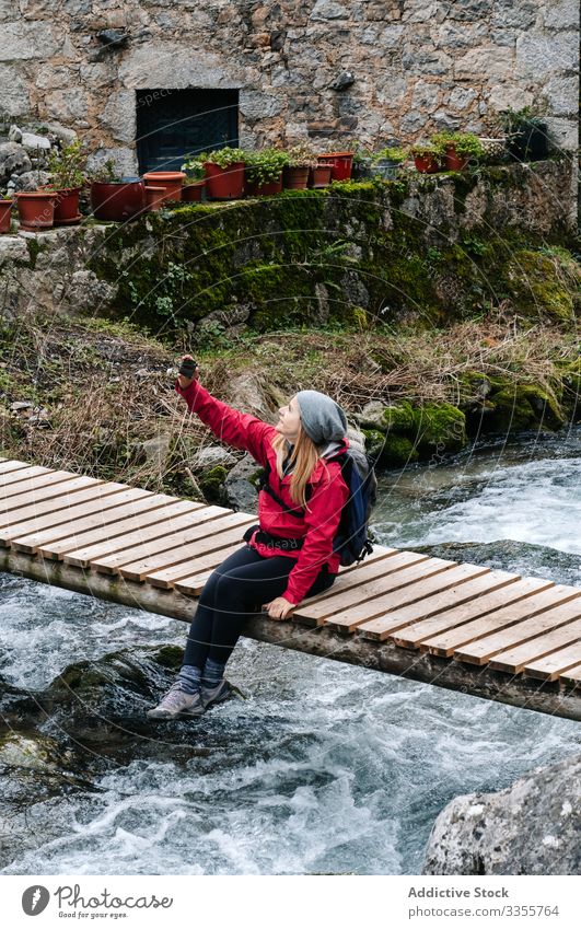 Pleased tourist with backpack taking selfie on smartphone sitting on bridge above mountain river village house using travel nature device gadget dry peak