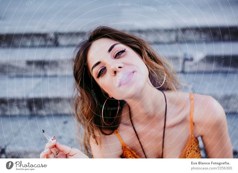 Beautiful young caucasian woman sitting on stairs at the city street on a sunny day and smoking a cigarette. Urban lifestyle and smoking concept. close up view