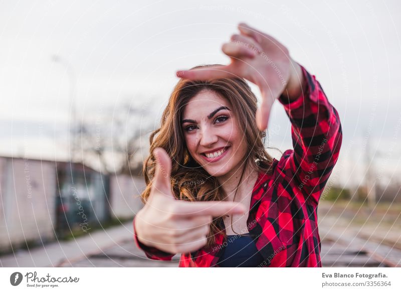 portrait of a young beautiful woman sitting on the floor and feeling happy. She is wearing casual clothes and making a frame with her hands.. Outdoors city background. Lifestyle