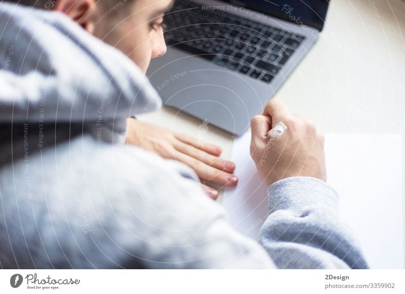 Back view of a young man writing on paper next to laptop at home Table School Study Academic studies Work and employment Profession Office work Workplace