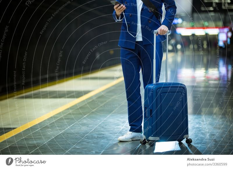 Young stylish handsome man in suit with suitcase standing on metro station holding smart phone in hand, scrolling and texting, smiling and laughing.  Train passing by