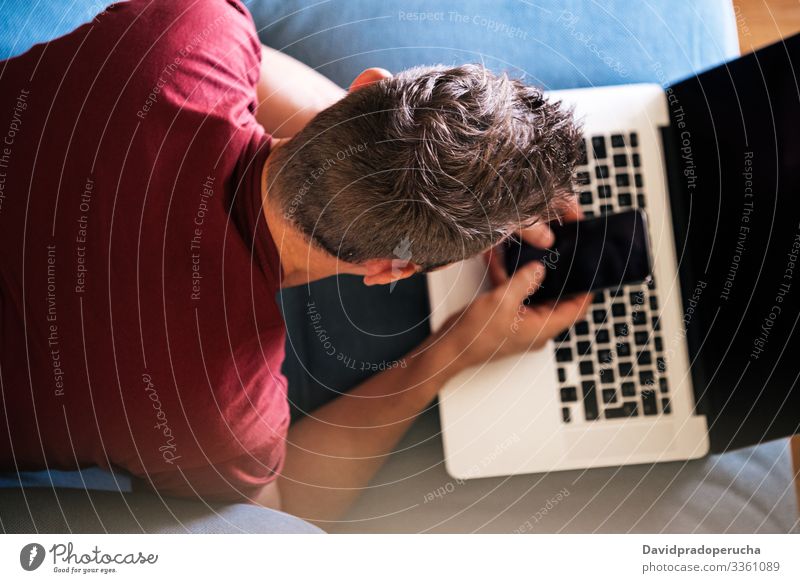 Casual male remote worker on sofa with laptop and smartphone man using couch lying freelance casual home browsing lounge typing keyboard connection