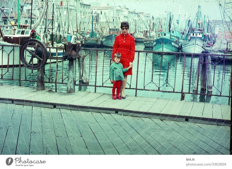 Happy, fashionable, stylish mother with daughter at hand, in partner look, are on holiday in the 60's, during a stroll through the city; trip on a bridge in the port of Venice, with water, ships and boats in the background.