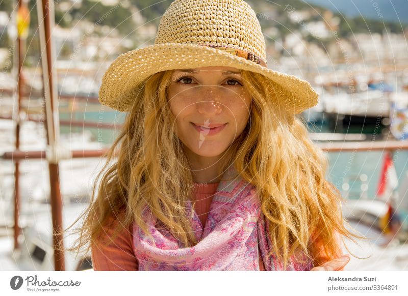 Portrait of young woman with light curly hair in straw hat enjoying sun and breeze, smiling. Sunny harbor with boats and yachts, green mountains on background. Enjoying life, happy person traveling,